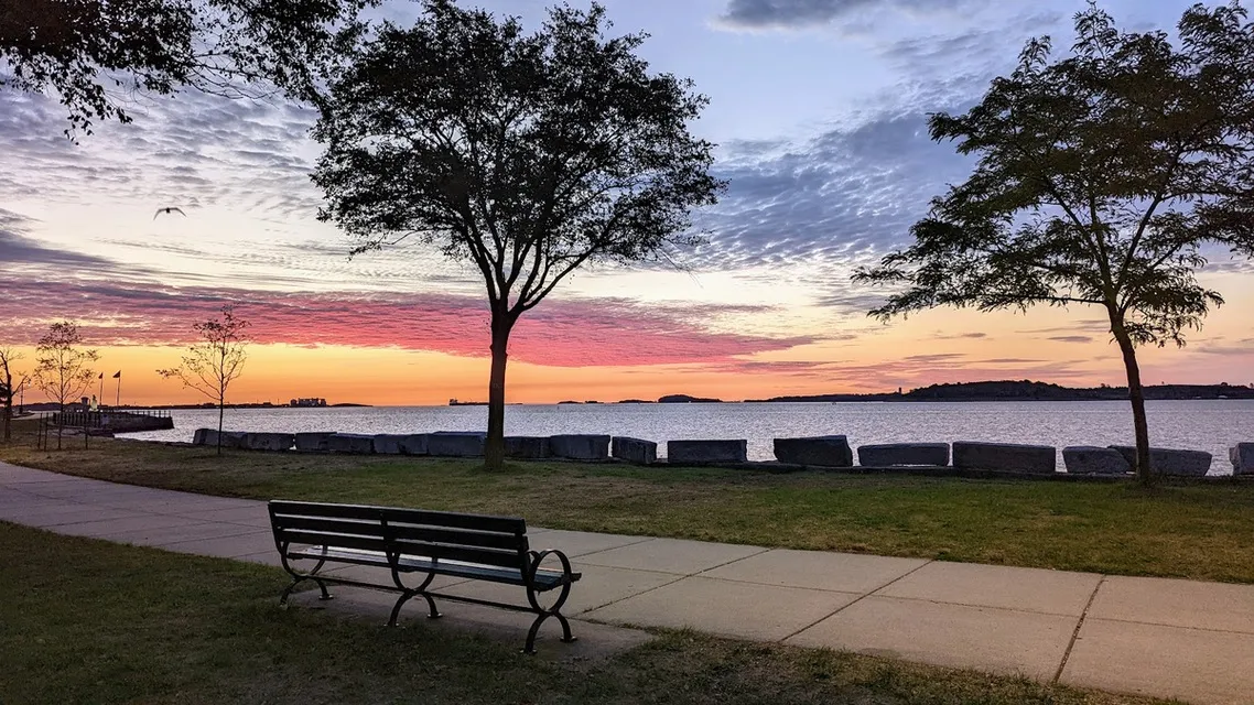 Park bench and harbor path at Castle Island under a sunset sky