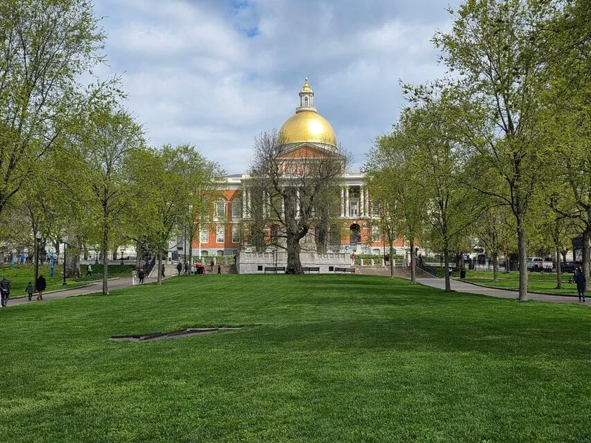 A lawn and trees on Boston Common leading to the gold dome of the Massachusetts State House on a spring day